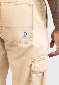 Beige cargobroek gemaakt van katoen; heeft meerdere zakken, een geweven label en zichtbare stiksels. De stof heeft een gladde textuur.
