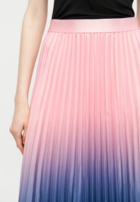 Femeie purtând o fustă plisată cu un degrade de la roz la albastru, asortată cu un top negru, mâna relaxată pe lângă corp.