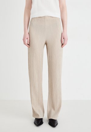 Beige, gestructureerde, wijde broek met hoge taille, gedragen met een wit mouwloos topje en zwarte puntige schoenen op een witte achtergrond.