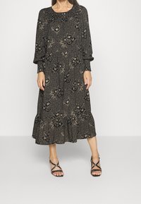 Frau, die ein schwarzes Midikleid mit beigen Blumen- und Polka-Dot-Mustern, langen Ärmeln und schwarzen, geflochtenen High Heels trägt, vor einem schlichten Hintergrund.