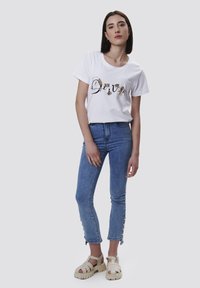 Camiseta corta blanca de manga corta con un gráfico de "Divino" en dorado y negro. Combinada con unos vaqueros ajustados de color azul claro y sandalias de tiras beige.
