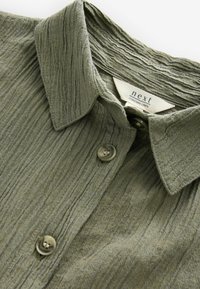 Olive green collared shirt with a textured fabric, featuring button closure and a label indicating it contains linen.