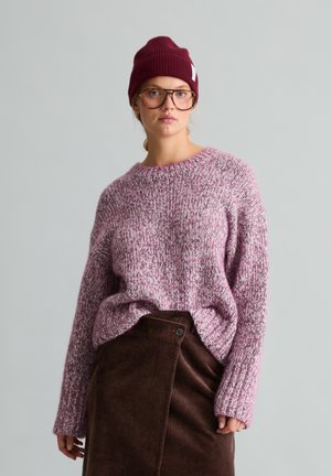Junge Frau trägt große Brille, bordeauxfarbenen Beanie, rosa-weißen Strickpullover und hoch taillierten dunkelbraunen Cordrock vor grauem Hintergrund.