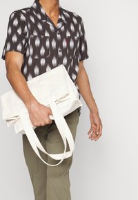 Uomo con camicia nera a maniche corte fantasia e pantaloni oliva che tiene una borsa tote color crema piegata per i manici.