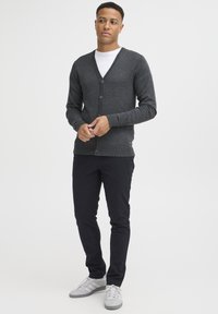 Mörkgrå cardigan med V-ringning, fem knappar, ribbade muddar, i kombination med svarta byxor och grå sneakers. Slät textur och figurnära passform.