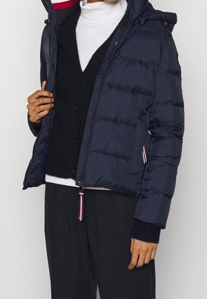 Persoon draagt een marineblauwe donsjas over een zwart vest met knopen en een witte coltrui, waarbij het jasje deels open is geritst.