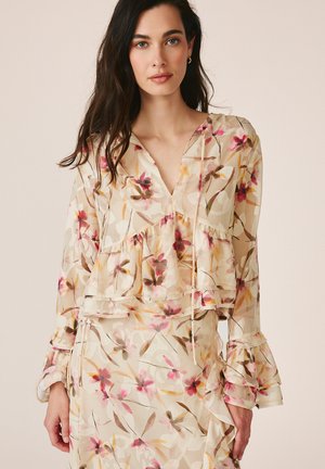 Mujer con cabello largo y oscuro que lleva un vestido beige con flores, volantes en capas y mangas largas sobre un fondo liso.