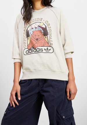 Femme portant un sweat-shirt beige avec une illustration d'un ours, des montagnes, des arbres et le texte "Official Guide Originals adidas", associé à un pantalon cargo sombre.