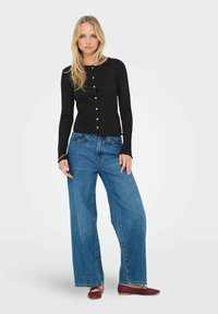 Czarny kardigan z prążkowanej tkaniny, okrągłym dekoltem i zapięciem na guziki, zestawiony z szerokimi niebieskimi dżinsami i czerwonymi balerinami. Gładka tekstura, casualowy krój.