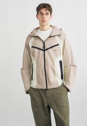 Ung mand iført beige og hvid jakke med lynlås og sorte lynlåse samt olivengrønne cargobukser, stående foran en ensfarvet grå baggrund.