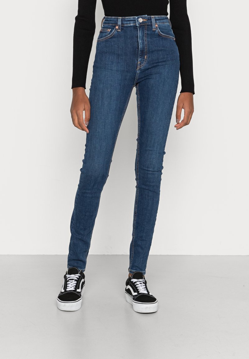 Personne portant un jean skinny bleu taille haute, un haut à manches longues noir et des baskets noires et blanches, debout devant un fond blanc.