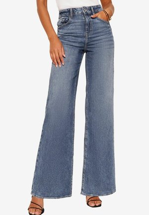 Hoch taillierte blaue Denim-Jeans mit weitem Bein, Kontrastnähten und Fronttaschen, präsentiert mit einem schlichten weißen Oberteil und Riemchensandalen.