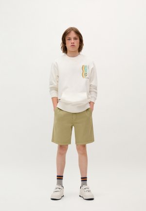 Sudadera blanca con un gráfico en el lado izquierdo, acentos verdes y naranjas, pantalones cortos caqui, zapatillas blancas con suelas negras y calcetines a rayas.