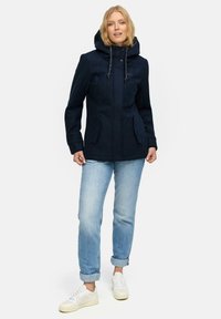 Femme portant une veste à capuche bleu marine, un jean clair retroussé et des sneakers blanches, se tenant devant un fond blanc uni.