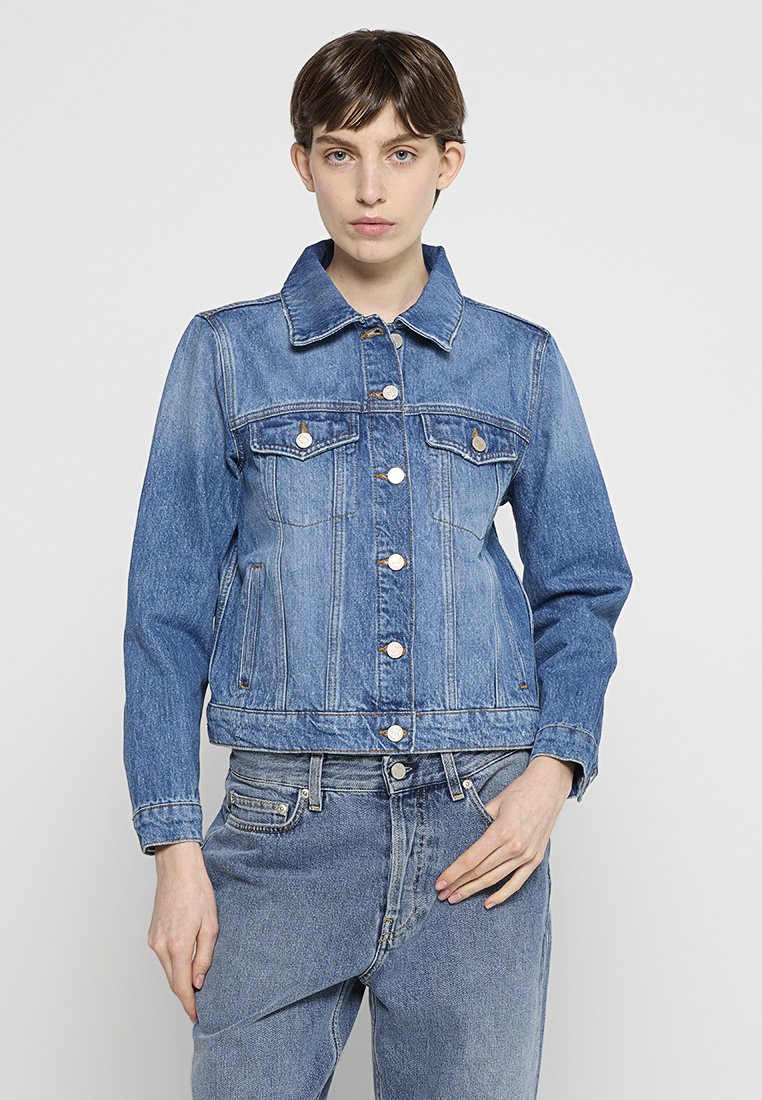 Junge Person mit kurzen braunen Haaren, die eine blaue Jeansjacke und blaue Jeans trägt, steht vor einem schlichten hellen Hintergrund.