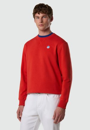 Jeune homme portant un sweat-shirt rouge avec un petit logo circulaire sur la poitrine et un pantalon blanc, sur un fond clair uni.