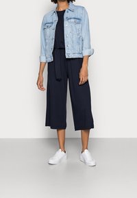 Jasnoniebieska kurtka dżinsowa, granatowy kombinezon w stylu culotte oraz białe trampki. Cechy obejmują związany w pasie krój, krótkie rękawy i luźny fit.