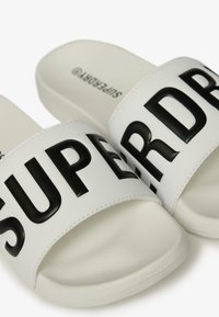 Infradito bianche con lettere nere larghe che leggono "SUPERDRY" sulle cinghie. Materiale testurizzato con una soletta liscia e piatta e una forma arrotondata.