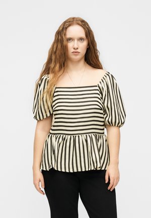 Femme aux longs cheveux roux ondulés portant un chemisier rayé vertical noir et blanc à col carré avec des manches bouffantes et un pantalon noir, debout devant un fond uni.