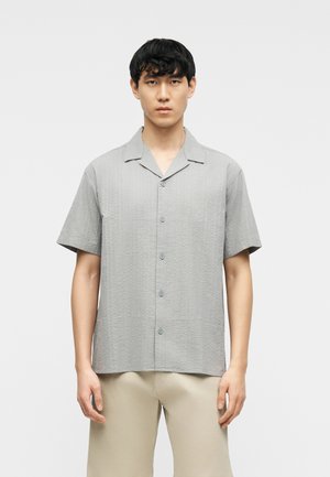 Jeune homme portant une chemise à manches courtes texturée gris clair avec des boutons et un pantalon beige, debout devant un fond blanc uni.