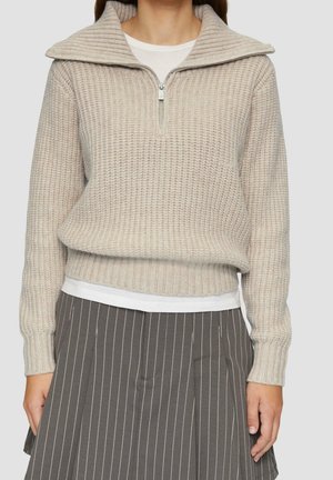 Femme portant un pull en maille gaufrée beige avec col demi-zip par-dessus une chemise blanche, assorti à une jupe plissée grise à fines rayures.