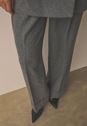 Pantalones gris de pierna ancha con bajos doblados combinados con zapatos negros de punta afilada sobre un suelo beige contra una pared clara.