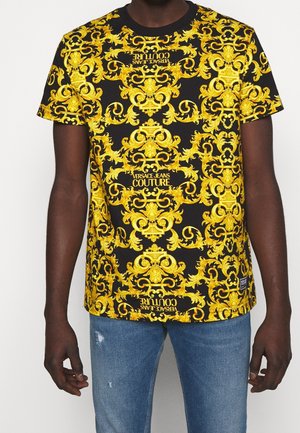 Hombre con camiseta negra con intrincados patrones barrocos amarillos y texto "Versace Jeans Couture", combinada con jeans azules desgastados.