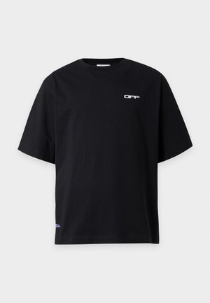 Homme debout portant un T-shirt noir oversize, un short gris clair, des chaussettes blanches et des baskets noires sur fond blanc.