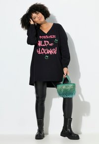 Abito nero a maniche lunghe con testo grafico rosa, leggings neri e una borsa verde con frange. La modella indossa stivali neri alla caviglia con borchie.