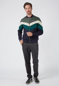 Chaqueta de hombre con cremallera y diseño de bloques de color en verde, azul marino, blanco y azul oscuro; fabricada en tejido suave con un dobladillo ajustado.