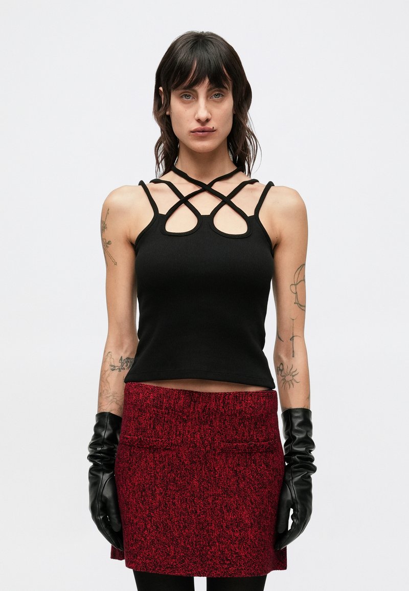 Zwart geribbeld topje met gekruiste bandjes, gecombineerd met een geruite rode rok. Model draagt zwarte handschoenen en toont een nauwsluitende taille.
