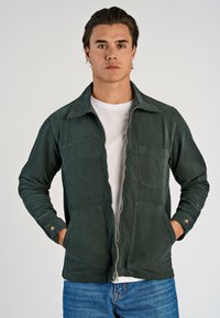 Groene corduroy jas met een ritssluiting aan de voorkant, spitse kraag, borstzak en twee zijzakken. Draagt over een witte T-shirt en blauwe jeans.