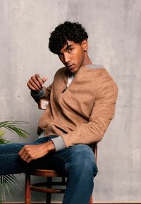 Veste bomber en daim marron avec des poignets tricotés gris, portée sur une chemise blanche. Jean bleu et chaise en bois, avec une plante verte au premier plan.