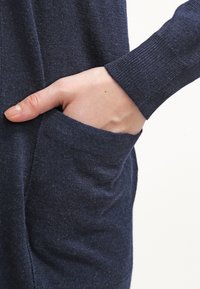 Námořnická modrá pletená látka zobrazuje ruku sahající do boční kapsy, přičemž ukazuje detail žebrovaného manžetu. Textura je měkká a lehce chlupatá.