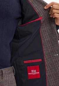Braun- und beige kariertes Blazer-Innenfutter mit schwarzem Futter, roten Akzenten und einem logo Patch. Enthält eine Tasche mit rotem Rand.