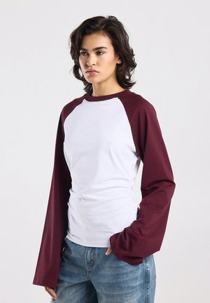 Jeune personne aux cheveux courts et ondulés, portant une chemise blanche à manches raglan rouge foncé et un jean bleu, debout devant un fond uni.