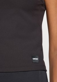 Camisola preta de alças com um patch do logótipo DKNY na parte inferior. Apresenta um design ajustado e uma textura suave. Costuras visíveis.