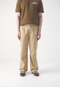 Beige cargo bukser med løs pasform, knaplukning og sidelommer, parret med en brun t-shirt og burgunderrøde sneakers.