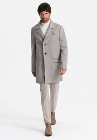 Abrigo de lana gris con grandes solapas, dos bolsillos frontales, cierre de un solo botón, combinado con pantalones beige y botas de ante marrón.