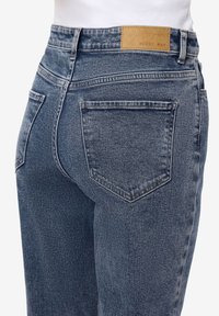 Høytlivde blå denimjeans med baklommer og en beige lærmerke med påskriften "NOISY MAY", brukt med en hvit topp.
