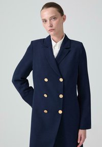 Marinblå dubbelknäppt kappa med guldfärgade knappar, skräddarsydd passform och slagkragar. Slät tyg med långa ärmar och en strukturerad design.