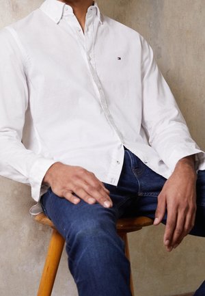 Mężczyzna siedzący na drewnianym stołku, ubrany w białą koszulę na guziki i niebieskie dżinsy, na tle beżowej ściany.