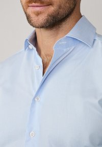 Lichtblauw gestreept overhemd met een puntige kraag, voorzien van witte knopen en een gladde textuur. Detail van het voorpand van het overhemd en het kraaggebied zichtbaar.