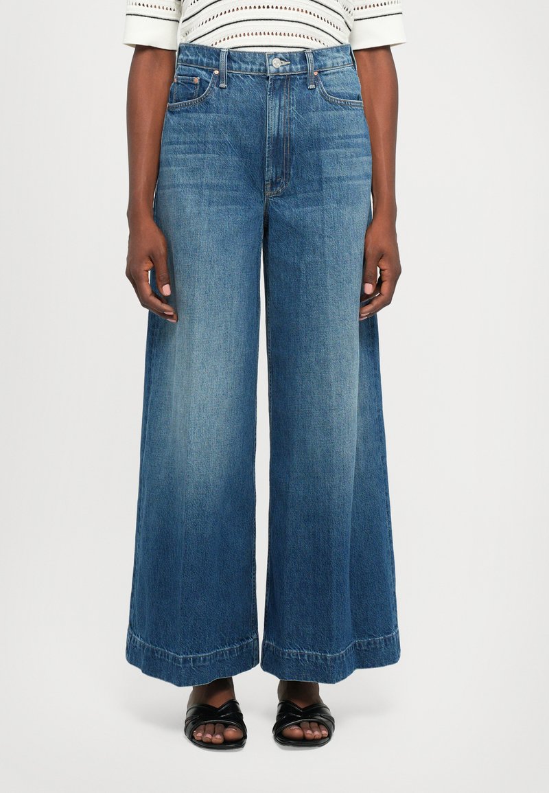 Personne portant un jean bleu taille haute à jambes larges avec des détails délavés, un haut blanc à manches courtes et des sandales noires à bout ouvert.