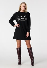 Robe sweat-shirt noire oversize à manches longues, avec le texte blanc "ICONIC STUDIOS". Portée avec des bottes en cuir bordeaux brillantes montantes jusqu'aux genoux.