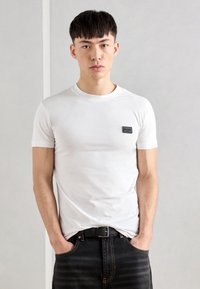 T-shirt bianca in cotone a maniche corte, colletto rotondo e un piccolo patch logo nero sul petto, abbinata a jeans di denim scuro.