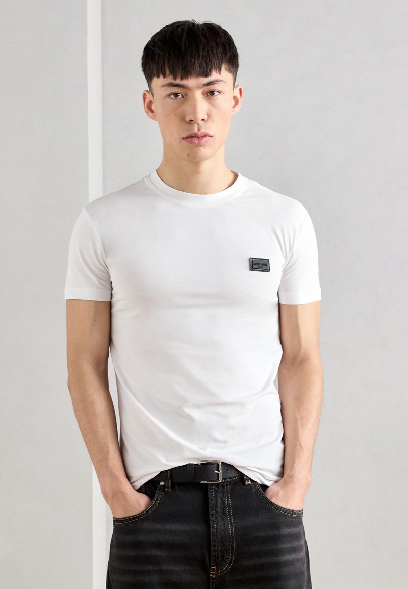 T-shirt bianca in cotone a maniche corte, colletto rotondo e un piccolo patch logo nero sul petto, abbinata a jeans di denim scuro.