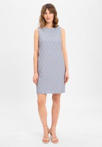 Robe sans manches de couleur bleu clair avec un motif géométrique doré. Coupe droite, mi-longueur, fabriquée dans un tissu lisse. Talons nus.