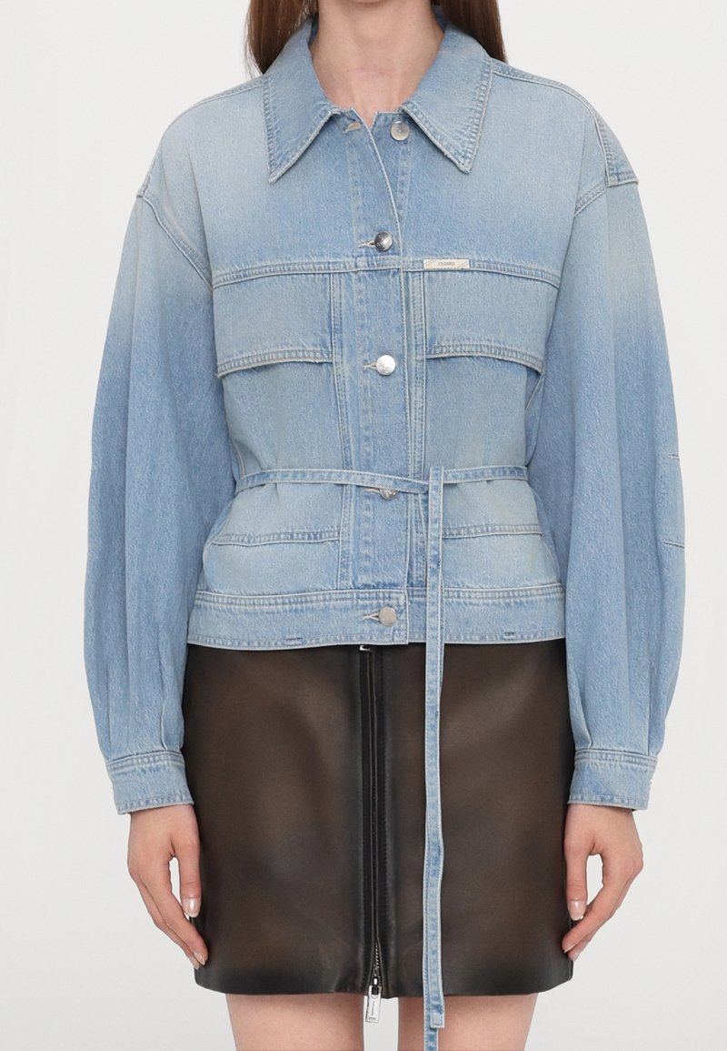 Veste en jean bleu clair avec large col, boutons argentés, ceinture et longues manches bouffantes portée sur une jupe en cuir noire avec fermeture éclair à l'avant.