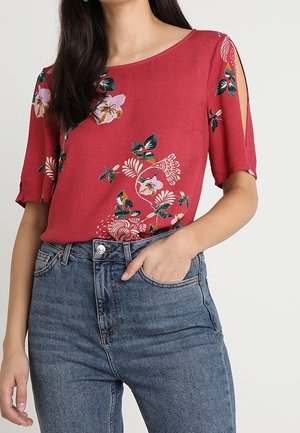 Vrouw draagt een rood gebloemde korte mouwen blouse met uitsparingen bij de schouders en high waist blauwe spijkerbroek, hand in de voorzak.